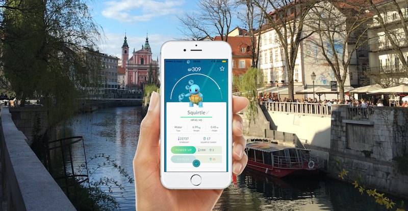 A hand holding a phone with the Pokemon GO app in the foreground and an image on the river in Ljubljana in the background.